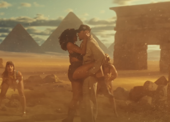 Chris Brown and a woman embracing passionately against a backdrop of the Egyptian Pyramids, with a third person observing them. The setting appears to be a dramatic, sand-covered landscape, possibly styled for a fantasy or historical film. The light suggests a sunset or sunrise, adding a warm tone to the scene.