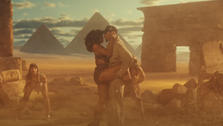 Chris Brown and a woman embracing passionately against a backdrop of the Egyptian Pyramids, with a third person observing them. The setting appears to be a dramatic, sand-covered landscape, possibly styled for a fantasy or historical film. The light suggests a sunset or sunrise, adding a warm tone to the scene.