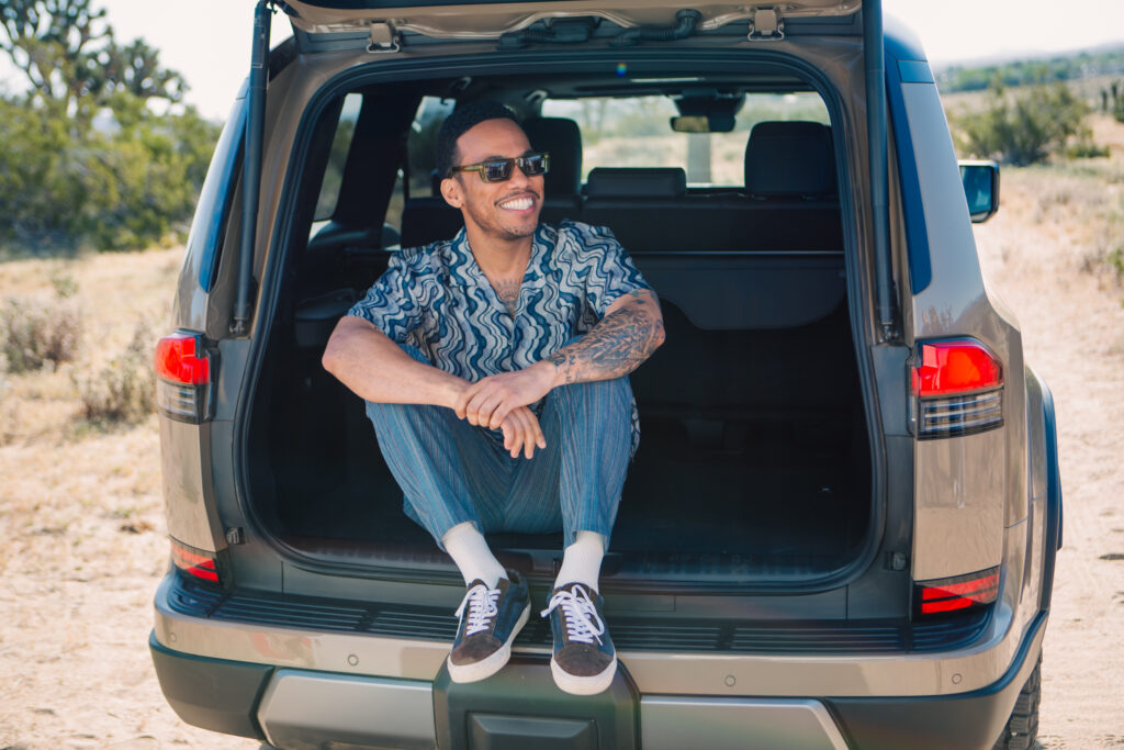 A photo of Anderson Paak wearing sunglasses and a patterned shirt sits smiling in the open trunk of a parked Lexus GX in a sunny, remote landscape.
