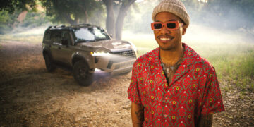 Anderson Paak wearing a patterned red shirt and sunglasses, paired with a beige hat, is smiling while standing outdoors. Behind him, a 2024 Lexus GX is parked amid a backdrop of trees illuminated by soft sunlight, with a mist of dust in the air