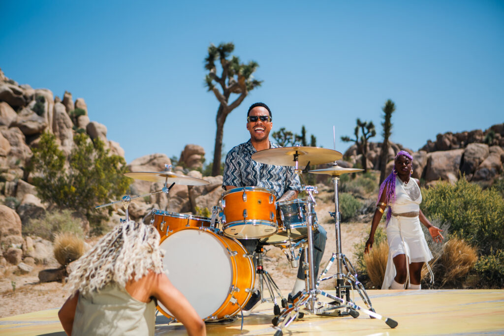Anderson Paak plays drums on a vibrant, decorated platform outdoors, surrounded by rocky terrain and scattered bushes under a clear blue sky. This photo is from a campaign with Lexus and Amazon Music