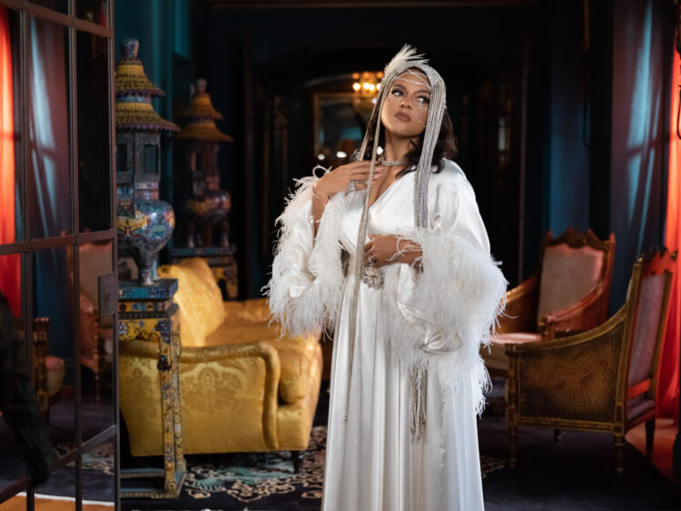 Marsha Ambrosius dressed in an elegant white outfit adorned with feathers and beads stands in a luxuriously decorated room, featuring rich blue walls and eclectic furnishings. Marsah Ambrosius is accessorized with a feathered headpiece and holds a thin, beaded accessory to their face. The ambient lighting gives the setting a warm, opulent atmosphere.